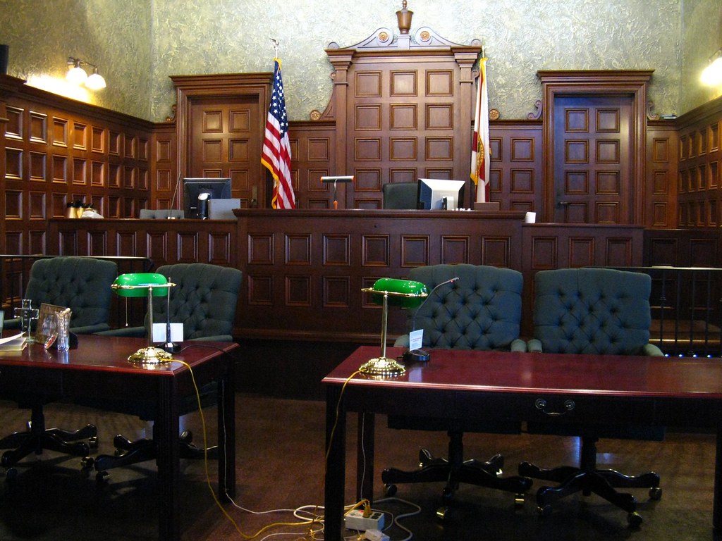 Image of a courtroom with 4 chairs behind two tables. An American flag and another flag in the background. Lots of wood detail from the doors to the walls as well as two green lamps on each table in the front. Who owns a Trademark Blog Post Image